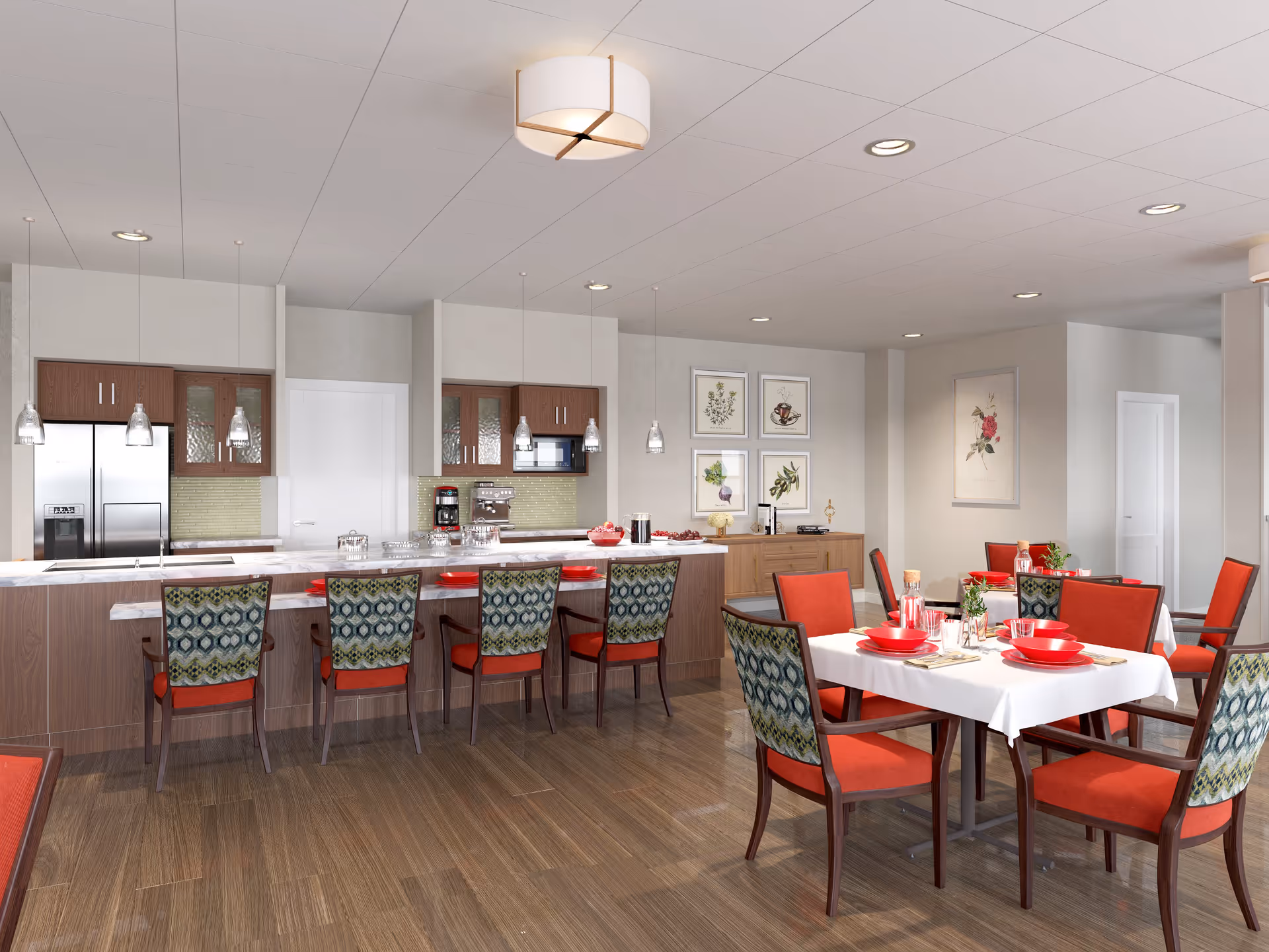 A modern dining area with a kitchen in the background. The kitchen features stainless steel appliances, wooden cabinets, and a marble countertop with pendant lights hanging above. In front of the kitchen counter are six chairs with patterned backs and red seats. To the right, there are two dining tables covered with white tablecloths, set with red plates, bowls, and cups, surrounded by red cushioned chairs. The room has wooden flooring and light-colored walls adorned with framed botanical artwork.