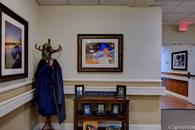 Interior hallway corner of a senior living facility with a wooden coat rack holding a blue jacket and a camouflage hat. A small wooden shelf below a framed picture on the wall displays several framed photos and a sign that reads 'UNITED WE STAND.' The hallway has beige walls with white trim and a carpeted floor, leading to another area with wood flooring and additional framed pictures on the walls.