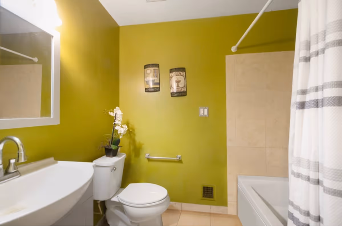 A bathroom with green walls featuring a white toilet with a potted orchid on top, a white sink with a silver faucet, a mirror above the sink, a bathtub with a white and gray striped shower curtain, and two decorative wall plaques.