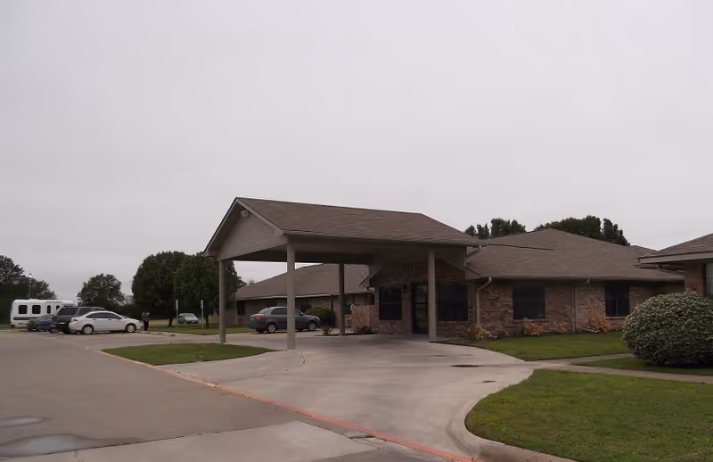 Brick senior living building with a covered drop-off entrance, driveway, parked cars, and trimmed lawns under an overcast sky.
