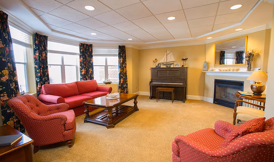 A cozy living room with a red sofa and two red armchairs with patterned upholstery. There is a wooden coffee table in the center, a piano against the far wall, and a fireplace with a mirror above it. The room has large windows with floral curtains and beige carpeted flooring.