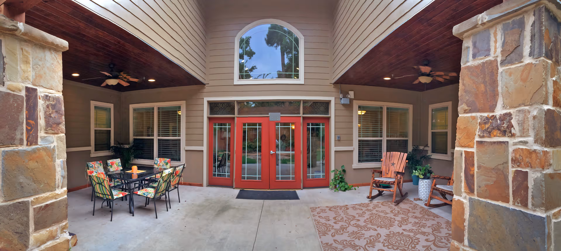 Covered outdoor patio area with stone pillars, a glass table with six floral cushioned chairs on the left, two wooden rocking chairs with cushions on the right, potted plants, ceiling fans, and red double doors with glass panels in the center.