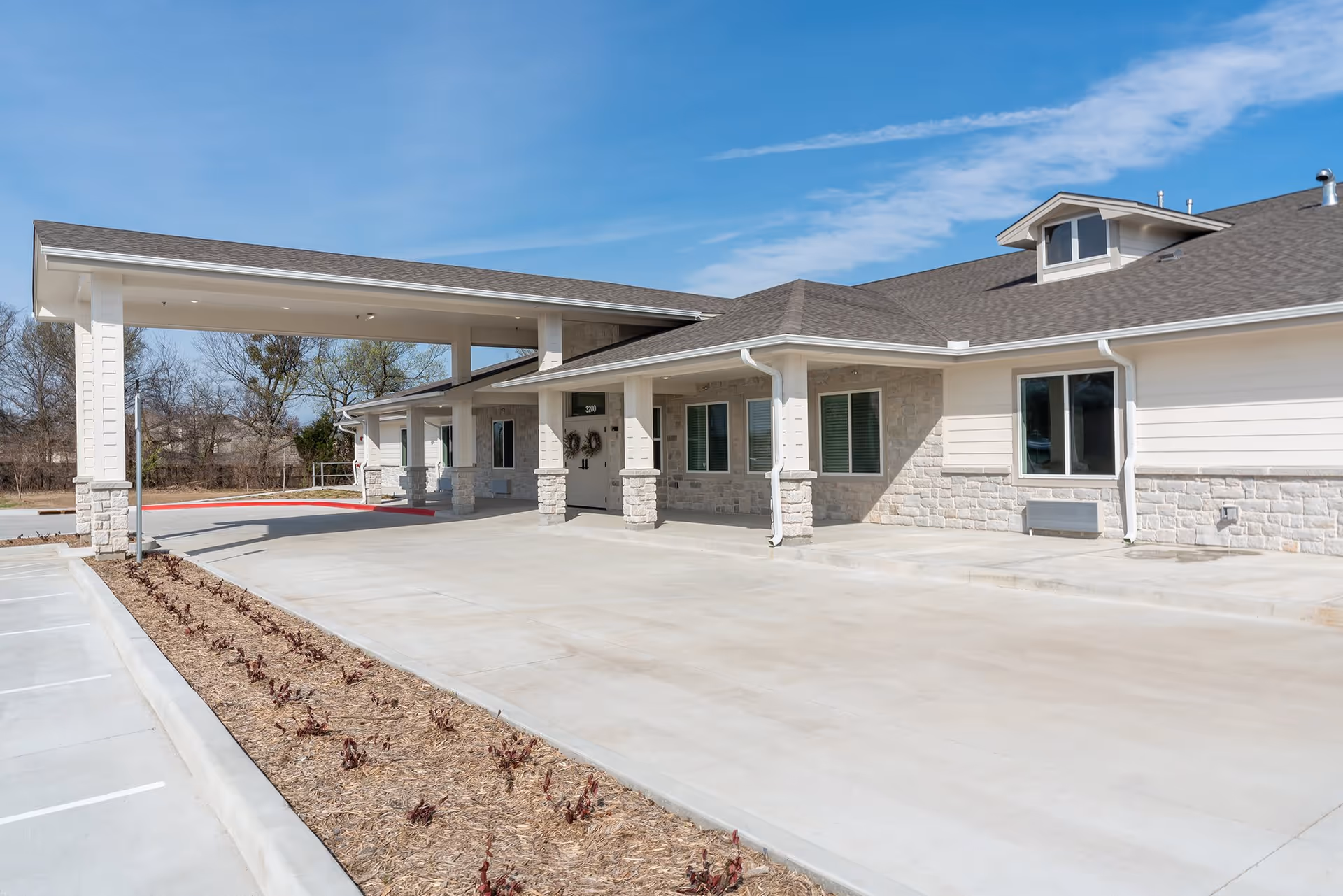Exterior view of a single-story senior living facility building with a covered entrance and stone and siding facade under a clear blue sky.