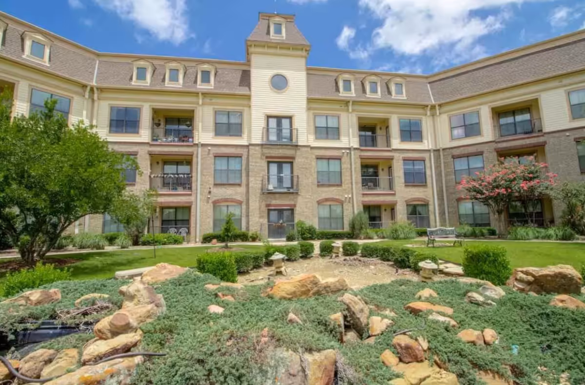 Courtyard with a landscaped rock garden and lawn in front of a three-story senior living building with balconies under a blue sky.