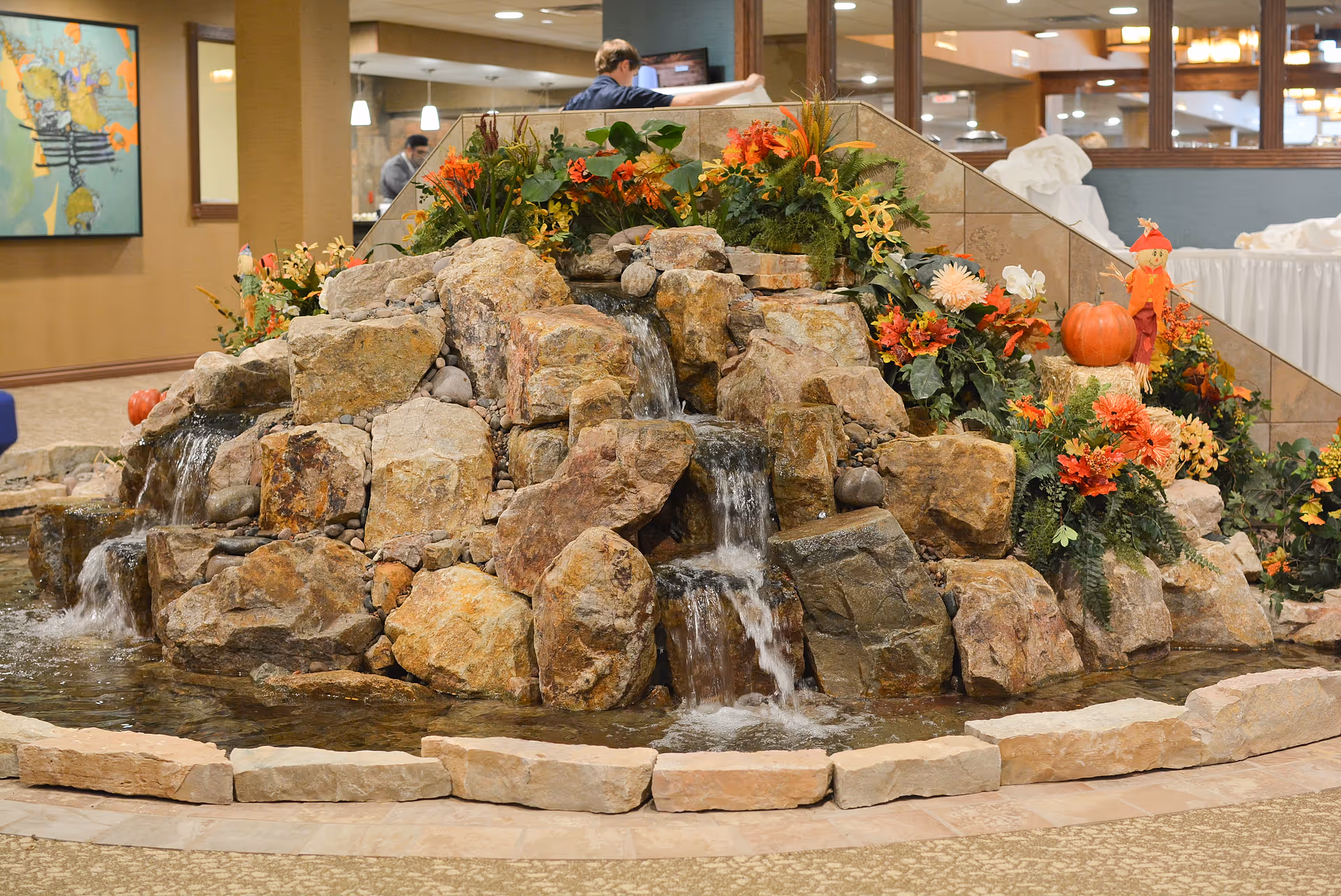 Indoor rock waterfall feature with flowing water surrounded by autumn-themed floral arrangements and decorations, including a pumpkin and a small scarecrow figure. The setting appears to be inside a building with carpeted floors and walls, and people are visible in the background.