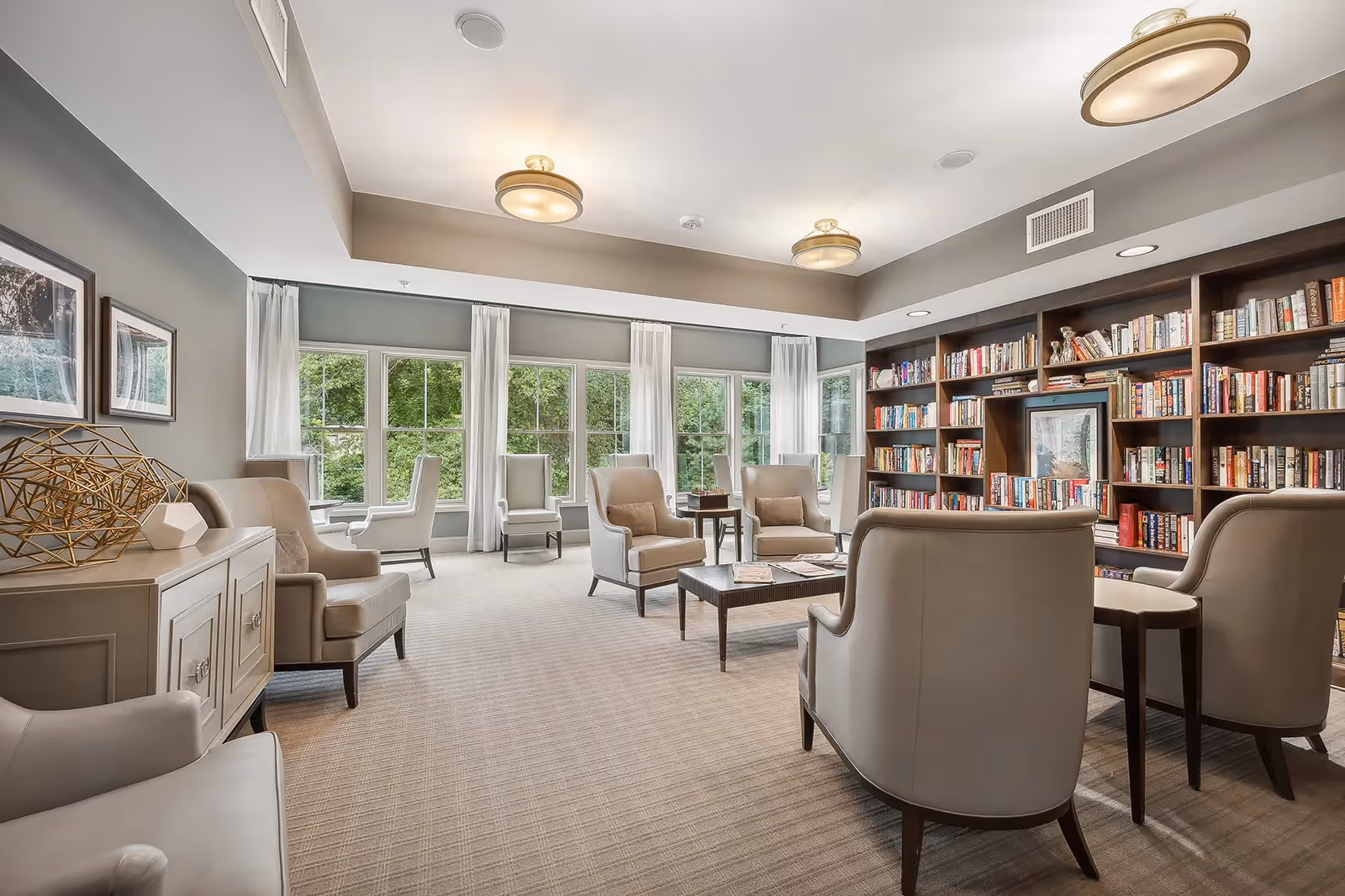 Bright communal library-style lounge with armchairs, coffee tables, large windows, and a wall of bookshelves.