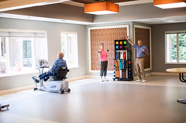 Three seniors exercising in a bright activity room — one on a recumbent exercise machine and two lifting small dumbbells near a rack of equipment.