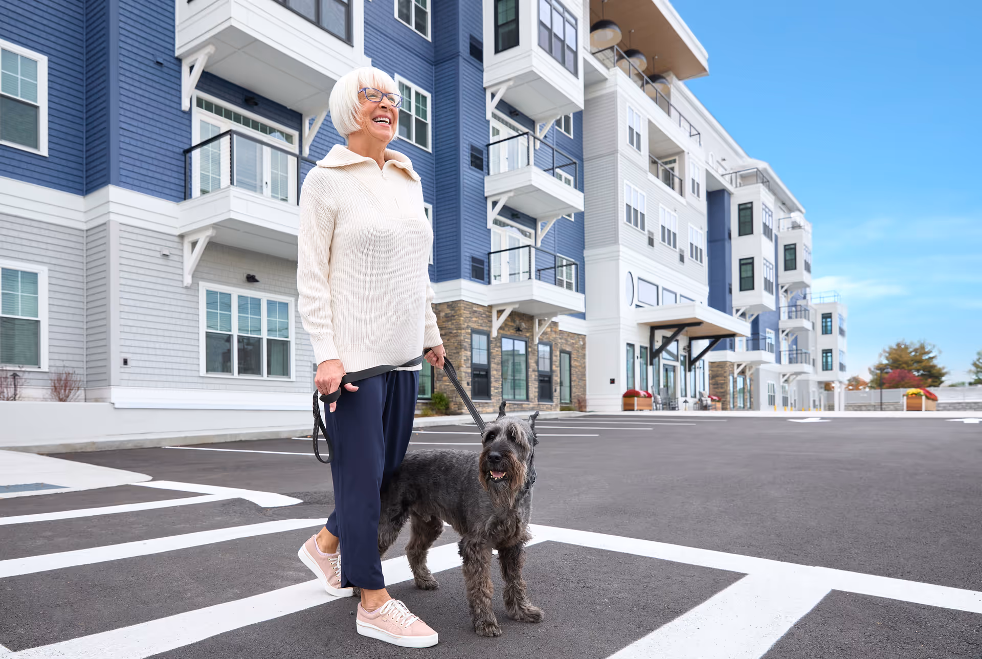 An elderly woman smiles while walking a dog in front of a multi-story senior living building.