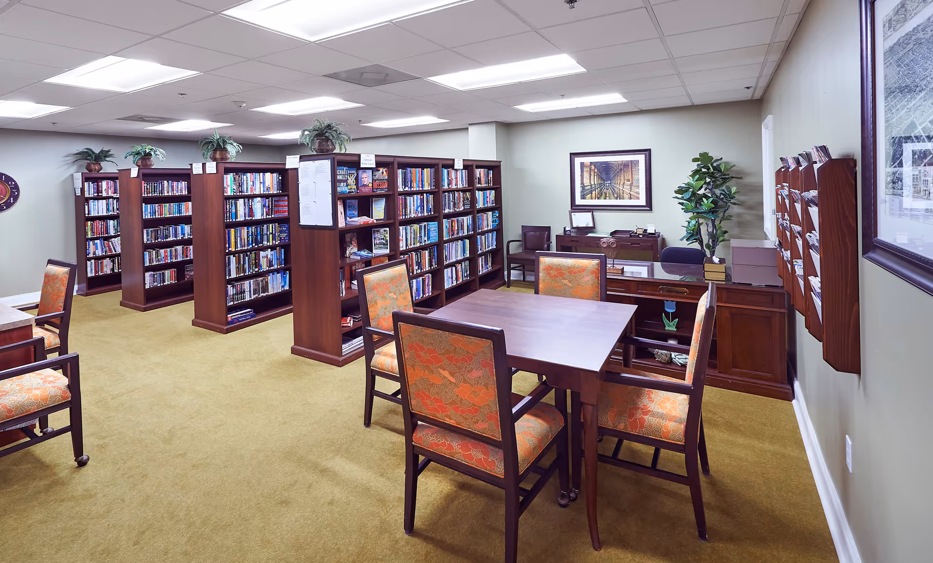 A quiet library room with multiple wooden bookshelves filled with books, a wooden table surrounded by four chairs with orange patterned cushions, and a desk with a chair and potted plant in the corner. The room has beige walls, carpeted floor, and framed artwork on the walls.