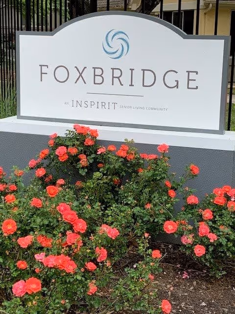 A white sign with the text 'FOXBRIDGE' and 'An INSPIRIT Senior Living Community' below it, mounted on a gray base. In front of the sign, there are blooming red and pink rose bushes. A black metal fence and part of a building are visible in the background.