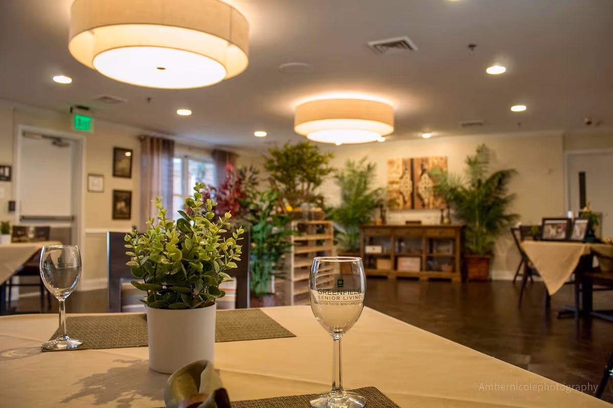 A dining room in a senior living facility with tables covered in beige tablecloths, each set with placemats, wine glasses, and small potted plants. The room is warmly lit with ceiling lights and decorated with various green plants and framed artwork on the walls.