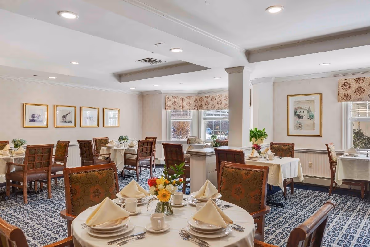 A well-lit dining room with multiple tables covered in white tablecloths, each set with plates, cups, silverware, and folded napkins. The room features patterned carpet, framed artwork on the walls, and windows with decorative valances letting in natural light. There are wooden chairs with cushioned seats around the tables, and a small floral centerpiece on the nearest table.