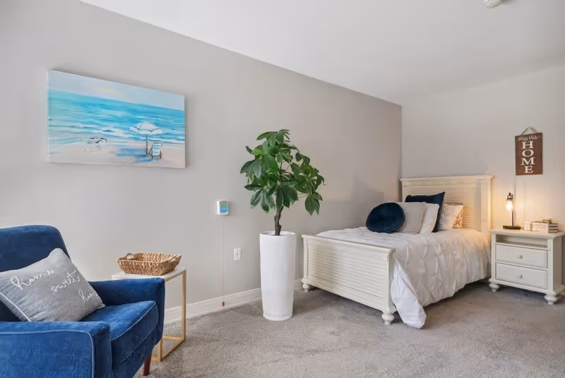 A cozy bedroom with a white wooden bed dressed in white bedding and blue accent pillows. Next to the bed is a white nightstand with a small lamp and books. A tall green plant in a white pot stands beside the bed. On the left side, there is a blue armchair with a gray pillow and a small side table with a wicker basket. A painting of a beach scene hangs on the light gray wall above the chair.