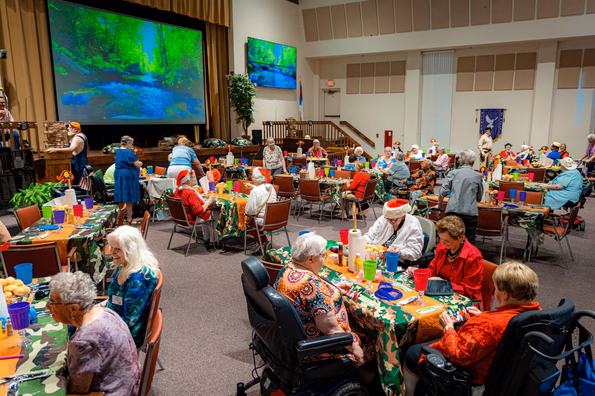 A large room filled with elderly people sitting at tables covered with camouflage-patterned tablecloths and orange runners. Many are engaged in conversation or activities, some wearing festive hats. The tables have colorful cups and plates, and there is a stage at the front with a large screen displaying a nature scene of a forest and river. The room has high ceilings and acoustic panels on the walls.