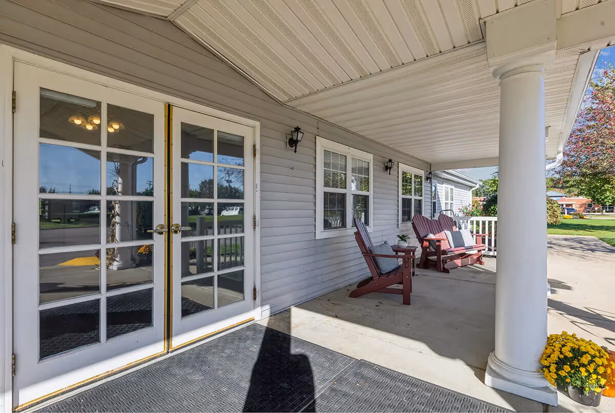 Covered outdoor porch area with white columns, two wooden rocking chairs with cushions, a small table with a plant, and double glass doors leading inside. There are windows on the building wall and a yellow flower pot near one of the columns.