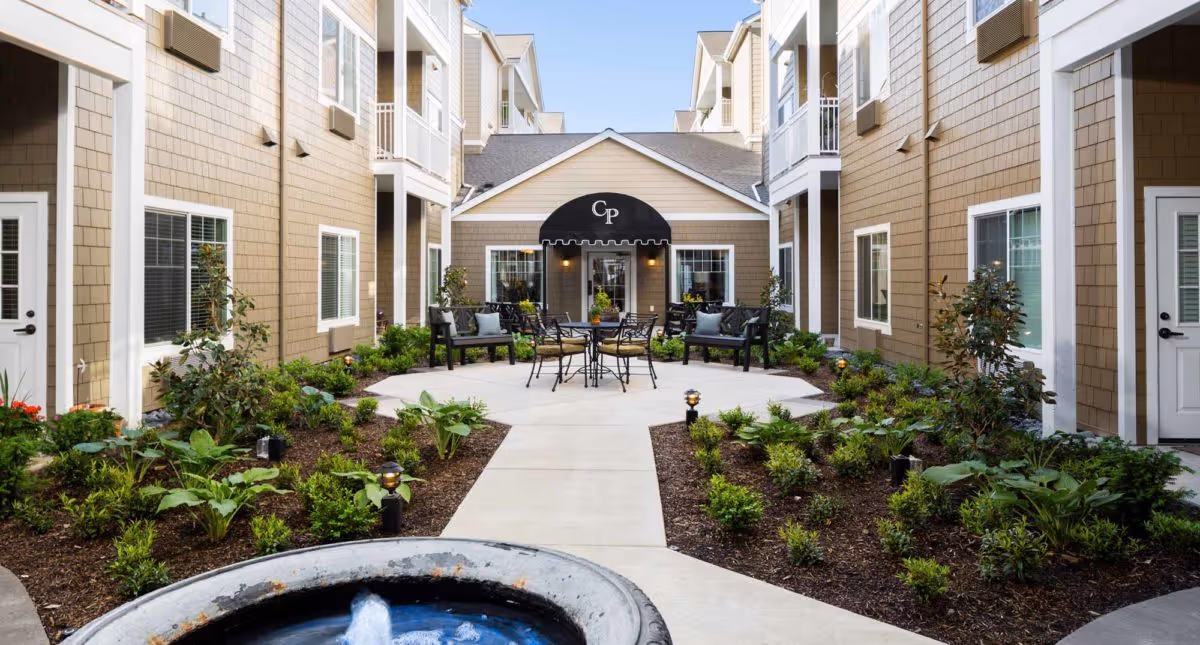 Outdoor courtyard area of Canterbury Park Independent Living Community featuring a central walkway, landscaped garden beds with green plants, a small water fountain in the foreground, and seating areas with chairs and tables near the building entrance with a black awning displaying the letters 'CP'.