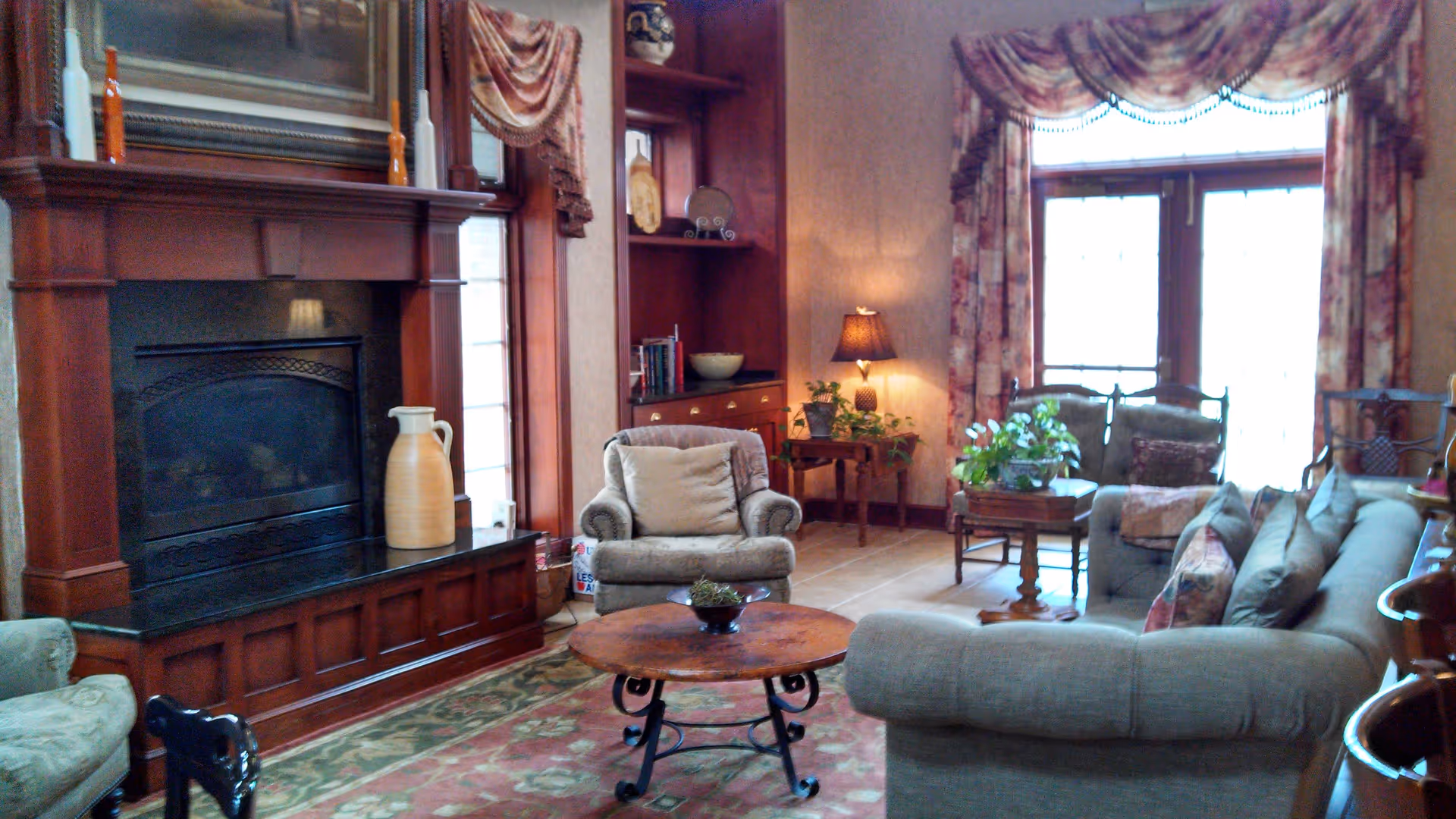A cozy living room with a wood-paneled fireplace, upholstered armchairs and sofa, coffee tables, plants, and large curtained windows.
