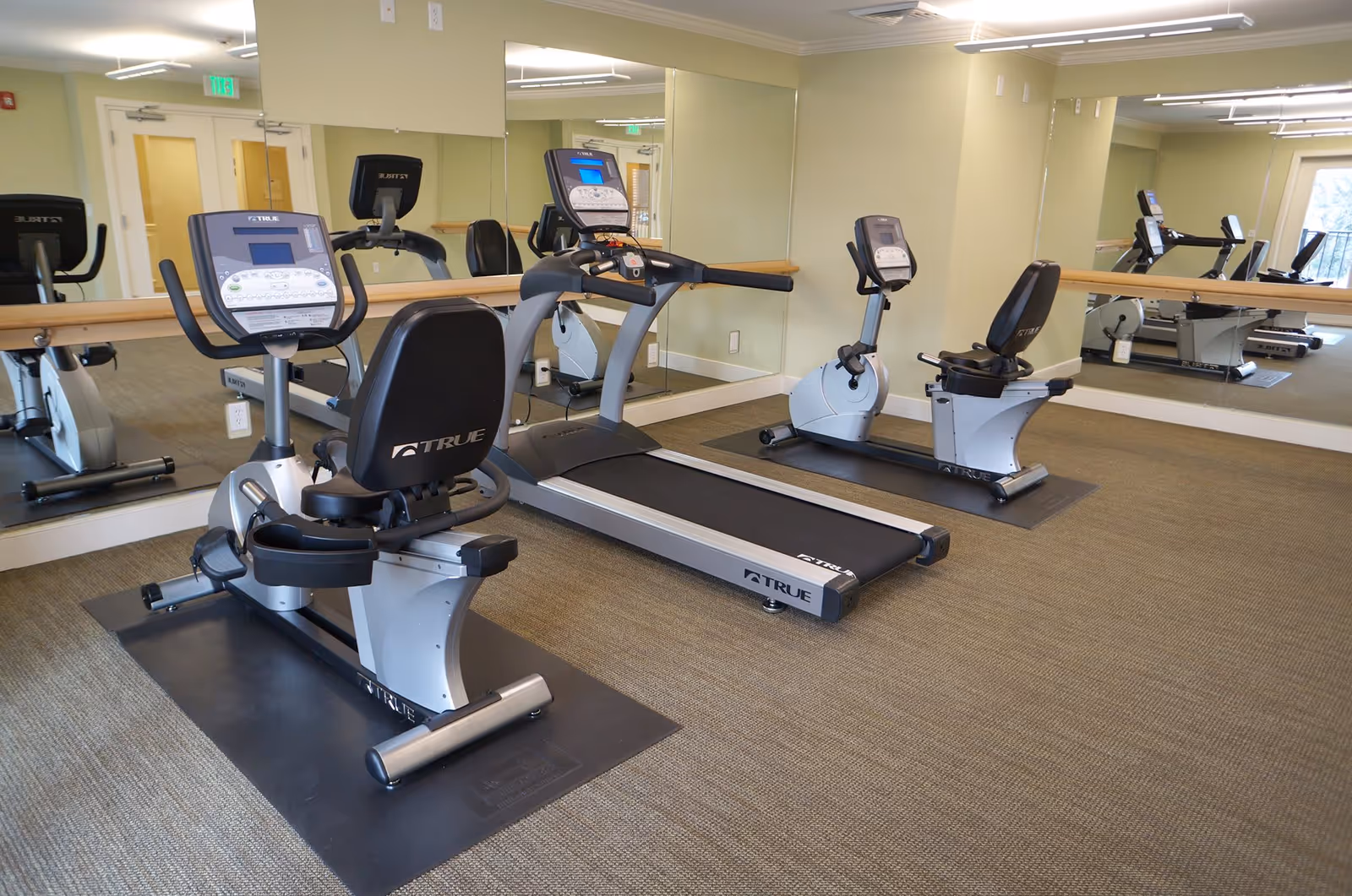 A small fitness room with exercise equipment including two recumbent stationary bikes and a treadmill. The room has beige walls, a large mirror covering one wall, and carpeted flooring. There is a door and an exit sign visible in the background.