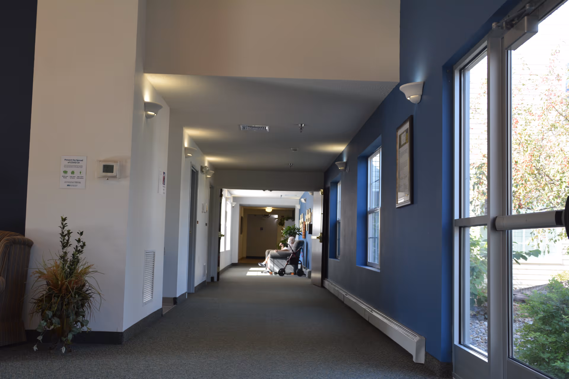 A long indoor hallway with blue and white walls, carpeted floor, and several wall-mounted lights. There are windows on the right side letting in natural light and a person sitting in a wheelchair near the end of the hallway. A potted plant is visible on the left side near a striped armchair.