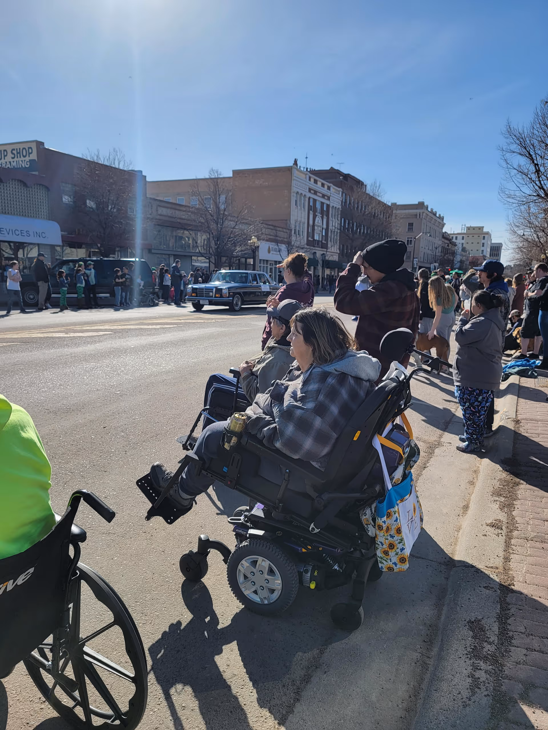 People gathered along a street on a sunny day, watching a parade or event. Two individuals in wheelchairs are in the foreground, with others standing or sitting nearby. Buildings and trees line the street in the background.