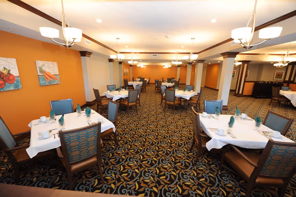A spacious dining room with multiple tables covered in white tablecloths, each set with green folded napkins, cups, and silverware. The room features patterned carpet flooring, orange accent walls with framed artwork, and modern ceiling light fixtures.