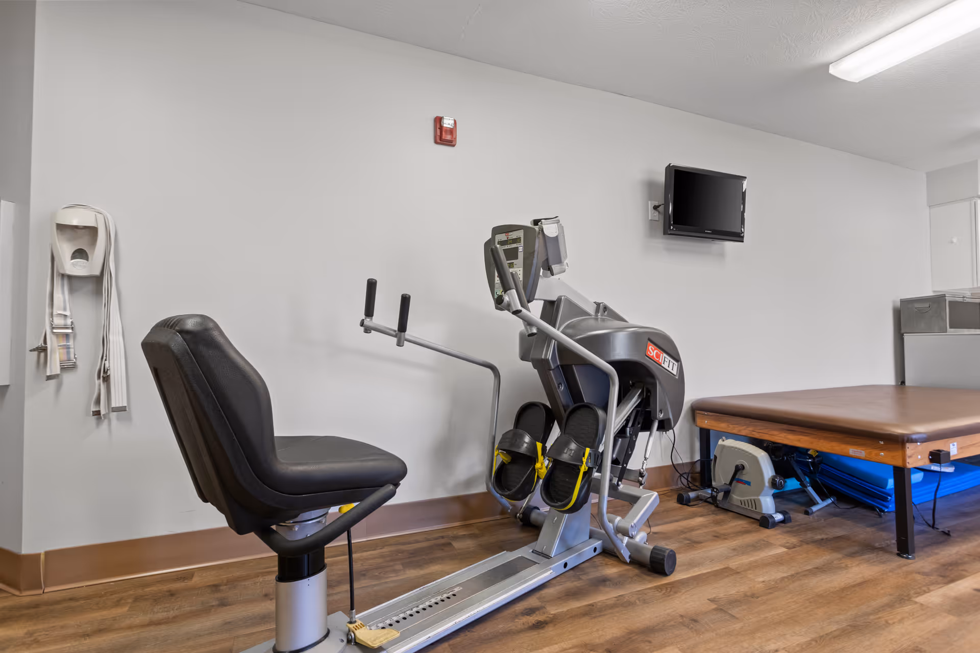 A small exercise room with a recumbent exercise bike labeled SCI FIT, a padded therapy table, a wall-mounted television, and a hand sanitizer dispenser on the wall. The room has wood flooring and plain white walls.