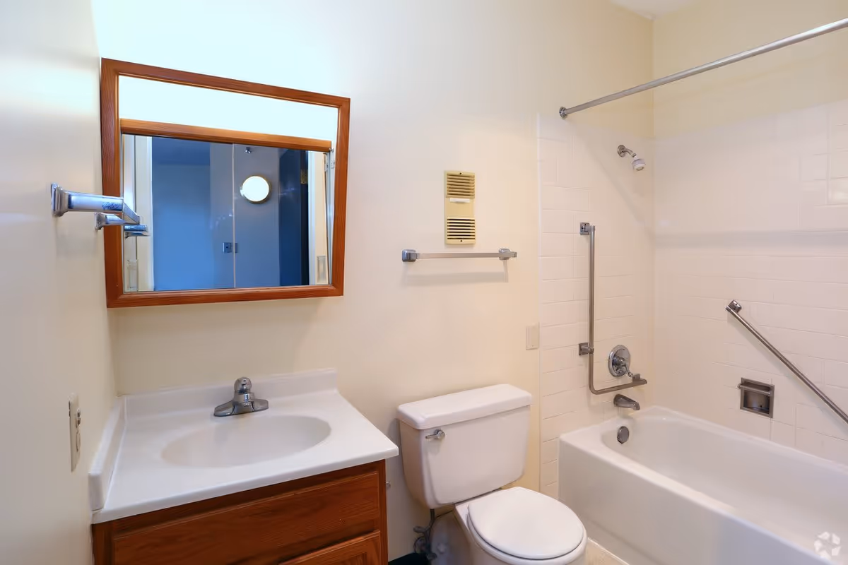 A clean bathroom with a white sink and wooden cabinet, a toilet, and a bathtub with white tiled walls and safety grab bars. A rectangular mirror with a wooden frame hangs above the sink.