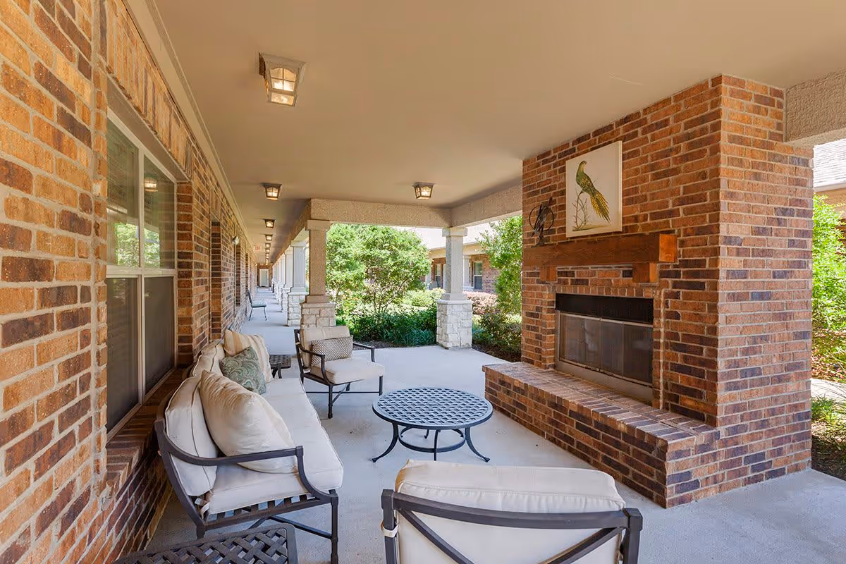 Covered outdoor seating area with cushioned chairs and a sofa arranged around a round metal table. There is a large brick fireplace with a wooden mantel and a framed bird artwork above it. The area is supported by columns and overlooks greenery and bushes.