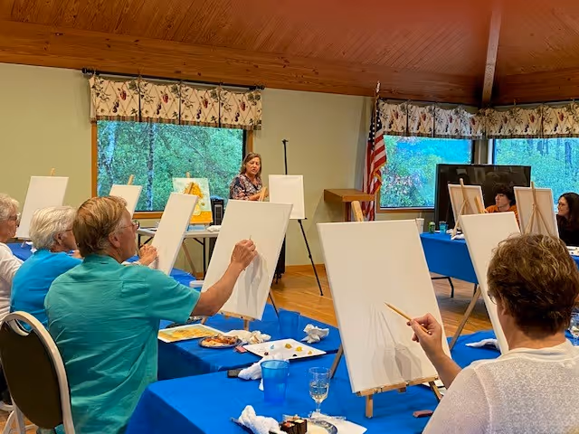 A group of elderly individuals seated at tables covered with blue tablecloths, participating in a painting class. Each person has a blank canvas on an easel and is holding a paintbrush. The room has large windows with floral curtains, wooden ceiling, and an American flag in the corner. A woman stands at the front of the room, presumably the instructor.