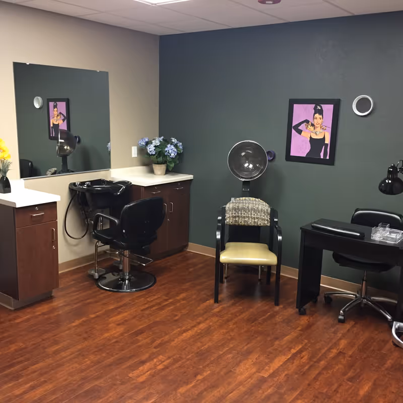 Interior view of a salon area with a hair washing station, a hair dryer chair, a manicure table with a lamp, and decorative flowers on a countertop. The walls are painted green and beige, and there is a framed artwork of a woman on the wall.