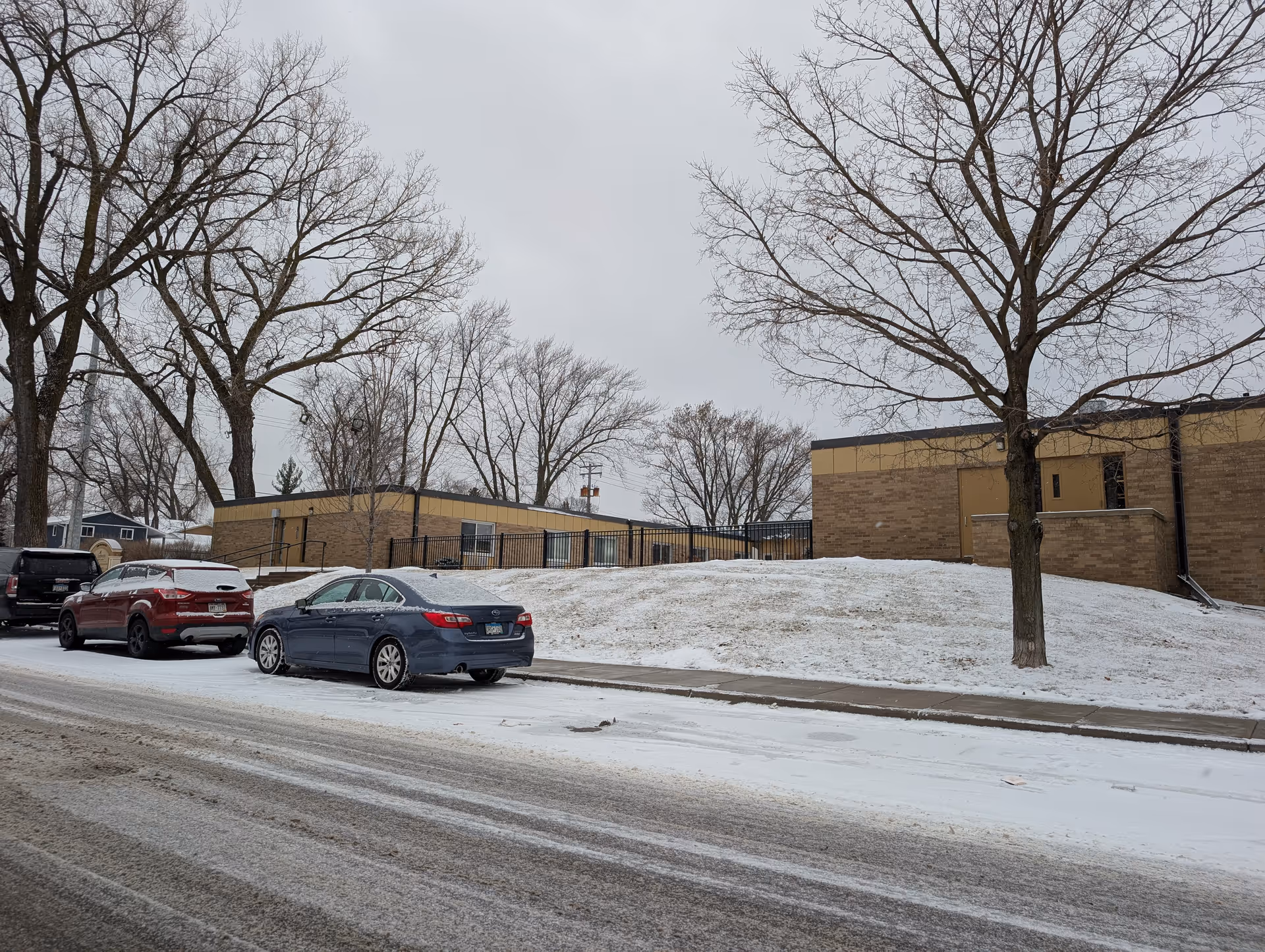 Snow-covered street with parked cars in front of a low brick building and leafless trees.