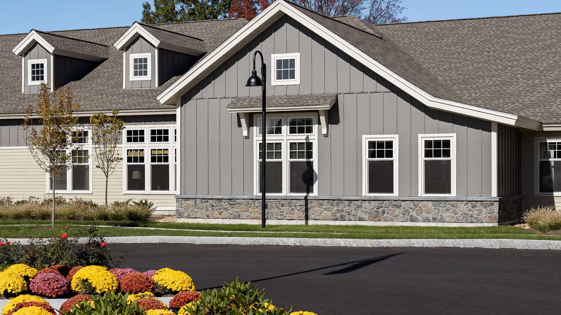 Exterior view of a senior living facility building with gray siding, white-trimmed windows, and a stone base. There are small trees and colorful flower beds in front of the building, along with a black street lamp casting a shadow on the wall.