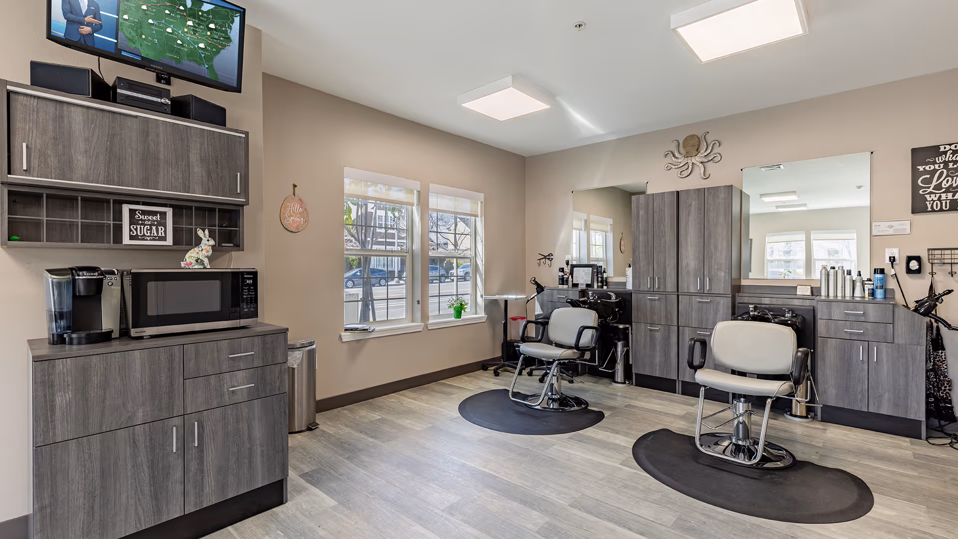 Interior view of a salon area in a senior living facility with two salon chairs on black mats, a large mirror, gray cabinets, a microwave, coffee maker, and a TV mounted on the wall showing a weather map. There are two windows letting in natural light and various decorative items on the walls and counters.