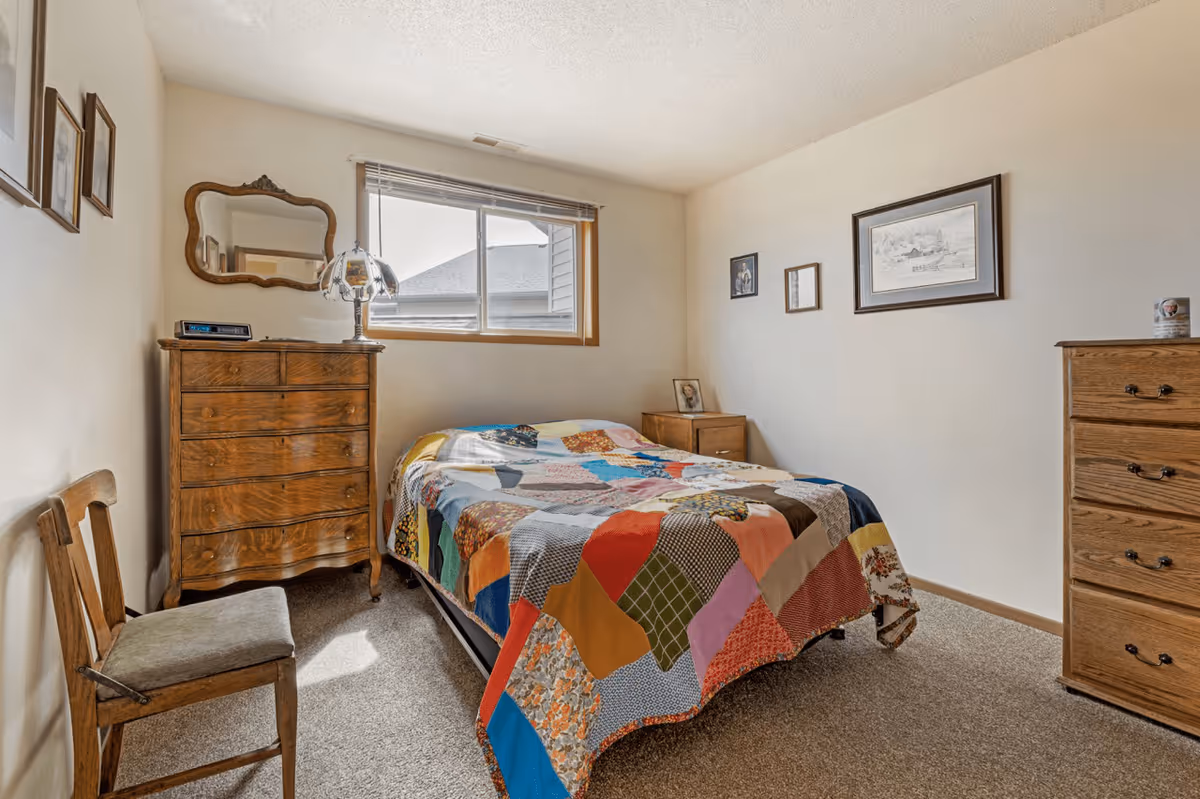 Small bedroom with a bed covered in a colorful patchwork quilt, wooden dressers, a chair, and a window.