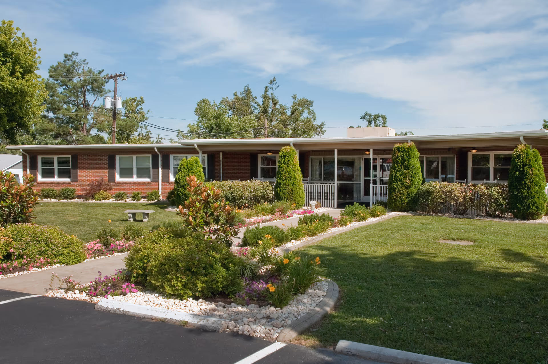 Single-story brick building with a covered entrance, surrounded by well-maintained green lawns, shrubs, and flowering plants under a partly cloudy sky.
