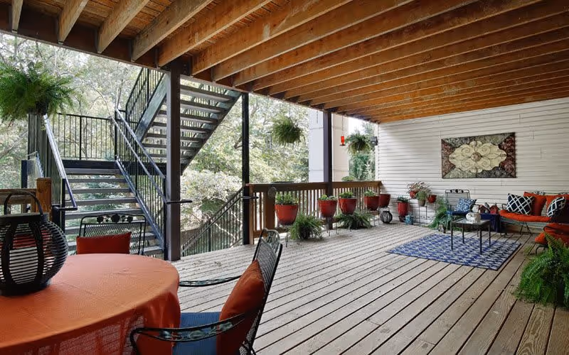 Covered outdoor patio area with wooden flooring and ceiling beams, featuring a round table with an orange tablecloth and black chairs with orange cushions. There are hanging plants, potted plants along the railing, a blue patterned rug, and a seating area with a red cushioned bench and pillows. Metal stairs lead down to a wooded area outside.