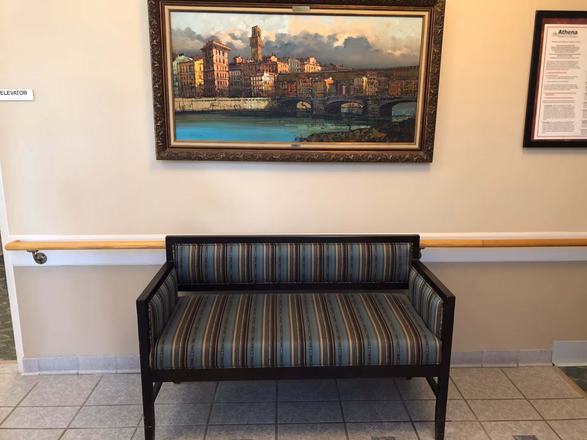 A small seating area with a striped upholstered bench against a beige wall. Above the bench hangs a framed painting depicting a cityscape with buildings and a bridge over water. To the left, a sign indicates the direction to the elevator. The floor is tiled and there is a wooden handrail along the wall.