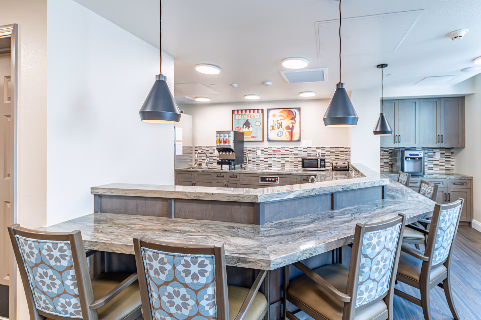 A modern kitchen area with a marble countertop bar surrounded by chairs with patterned backs. The kitchen features gray cabinets, a tiled backsplash, and appliances including a microwave and a coffee machine. Two pendant lights hang above the bar, and there are framed ice cream-themed artworks on the wall.