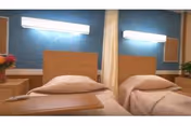 A room with two single beds separated by a curtain. Each bed has a wooden headboard, white bedding, and a pillow. There are wall-mounted lights above each bed and small bedside tables with flowers on either side.
