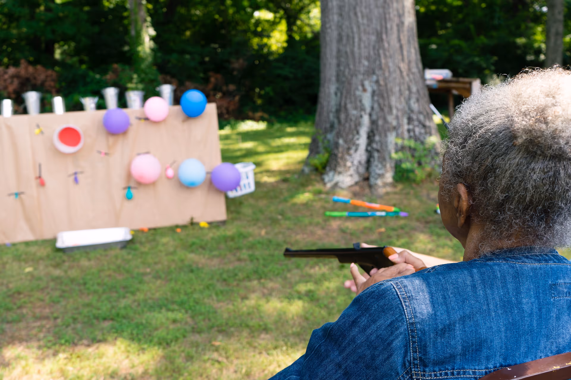 An elderly person with gray hair wearing a blue denim jacket is aiming a toy rifle at colorful balloons attached to a board outdoors in a grassy area with trees in the background.