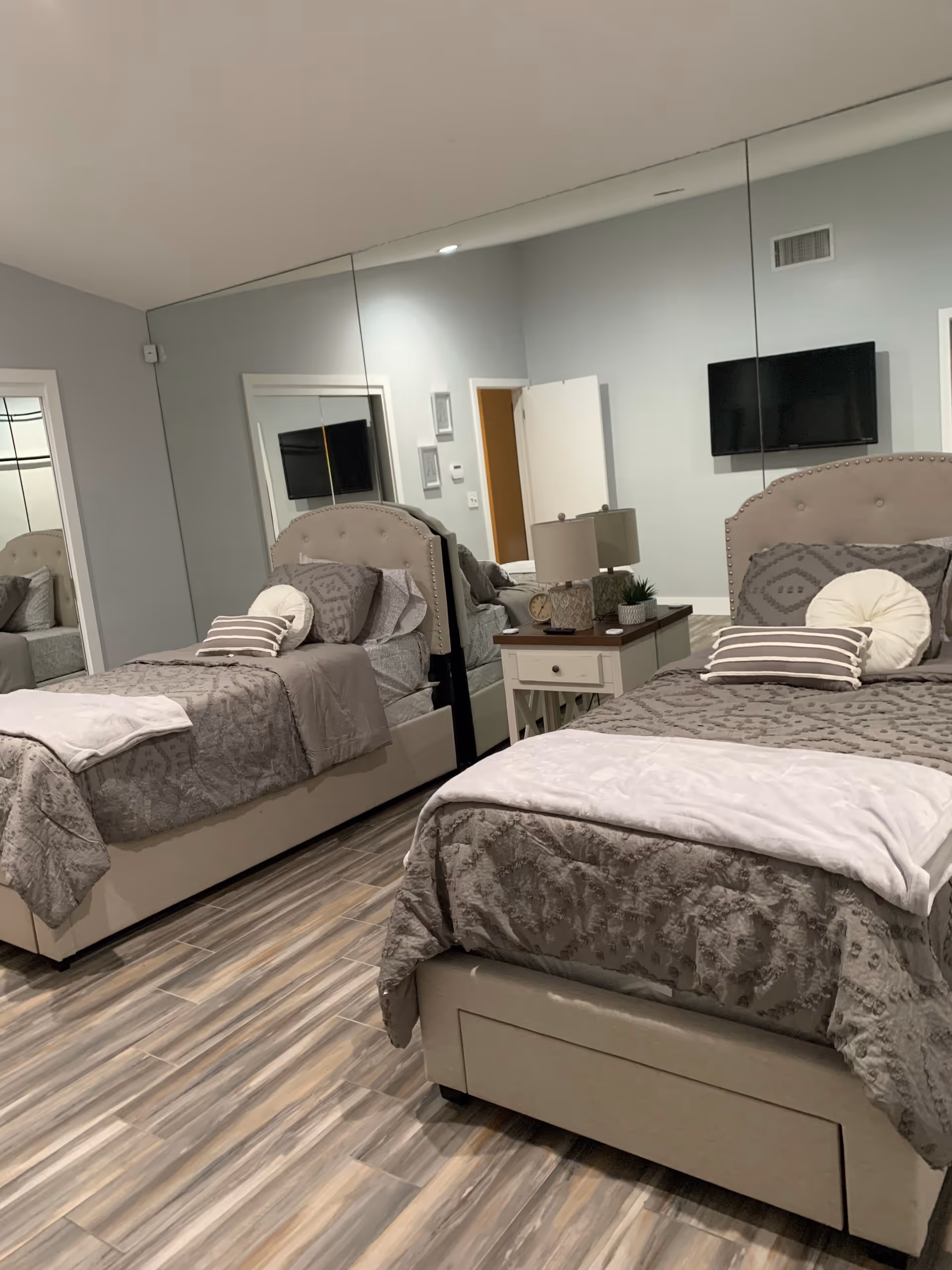 A mirrored bedroom with two twin beds, a central nightstand with a lamp and decor, and a wall-mounted TV.