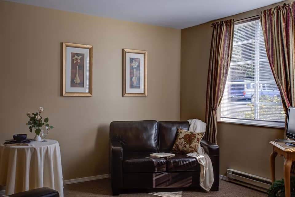 A cozy living room area with a dark brown leather loveseat adorned with a floral pillow and a white throw blanket. Next to the loveseat is a small round table covered with a white tablecloth, holding a vase with flowers, a glass, and a bowl. Two framed floral artworks hang on the beige wall above the loveseat. A window with striped curtains allows natural light to enter the room, and a small wooden table with a TV is visible on the right side.