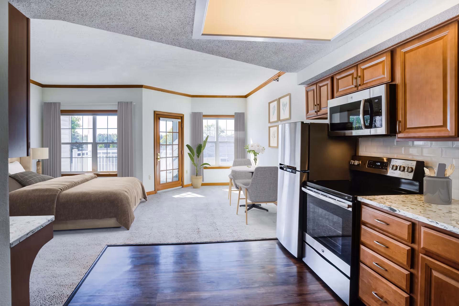 A bright and spacious studio apartment featuring a kitchen area with wooden cabinets, stainless steel refrigerator, stove, and microwave. The living and sleeping area includes a bed with brown bedding, a small dining table with two gray chairs, large windows with gray curtains, a glass door leading outside, and a potted plant near the corner.