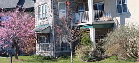 Exterior view of a residential building at Pennswood Village with stone and beige siding, featuring multiple windows and a balcony. The surrounding area includes green grass, bushes, and trees with pink and red blossoms.
