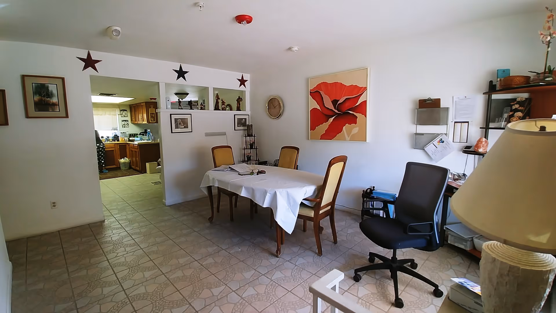 Interior view of a dining room with a rectangular table covered with a white tablecloth and four chairs around it. The room has tiled flooring and white walls decorated with framed pictures and a large red flower painting. There is a black office chair, a lamp, and a shelving unit with various items on the right side. An open doorway leads to a kitchen area with wooden cabinets and a person partially visible.