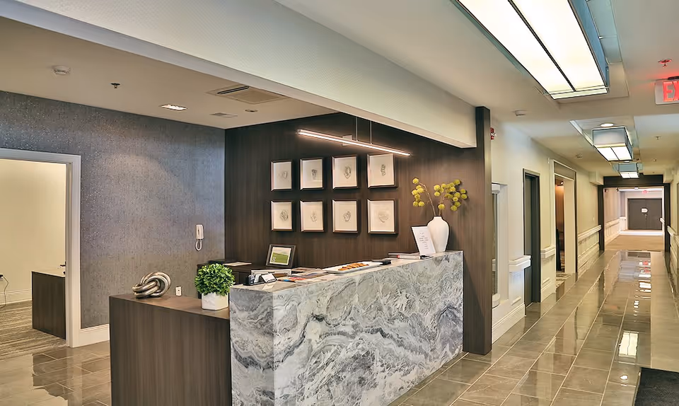 Reception area in The Parkdale Senior Living facility featuring a marble front desk with decorative items, framed artwork on a dark wood wall, and a long hallway with doors and overhead lighting.