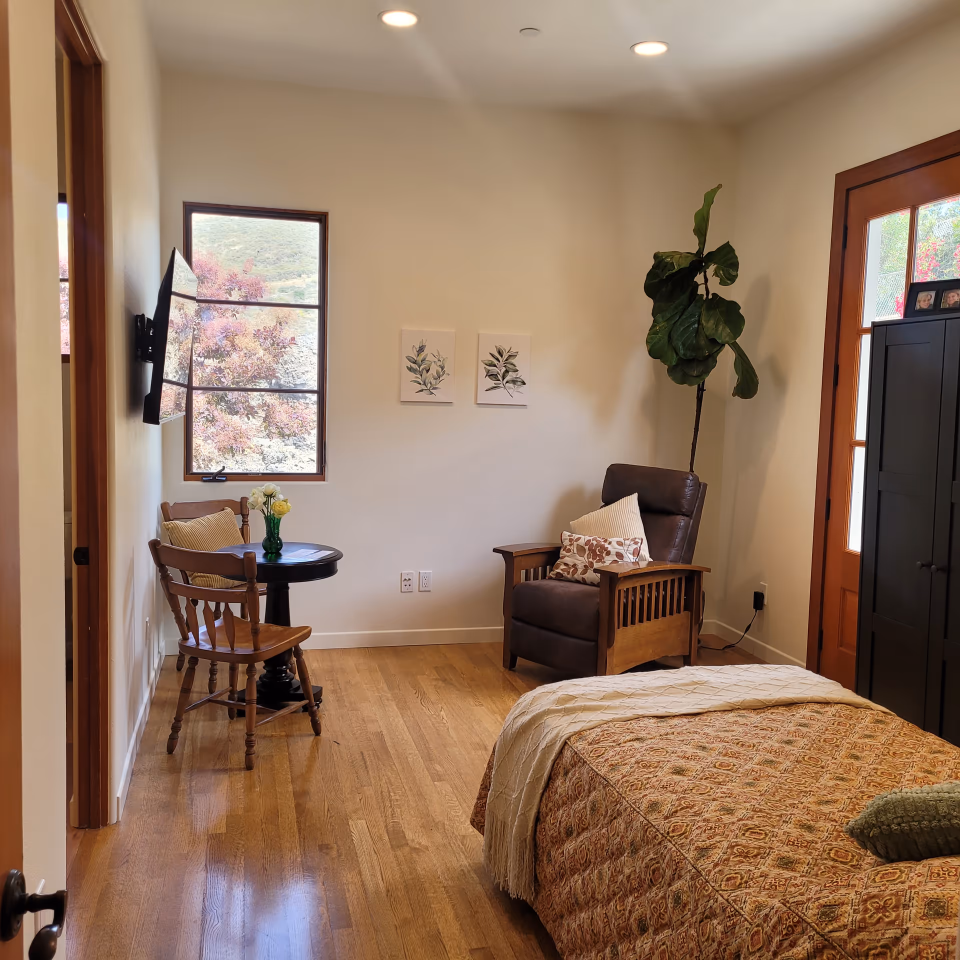 Sunny bedroom with a bed in the foreground, a wooden armchair and throw pillows, a small round table with chairs by a window, and a tall potted plant near the door.