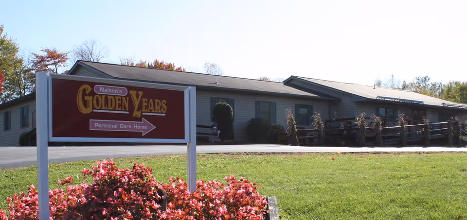 Exterior view of Nelson's Golden Years personal care home building with a sign in the foreground surrounded by pink flowers and green grass under a clear sky.