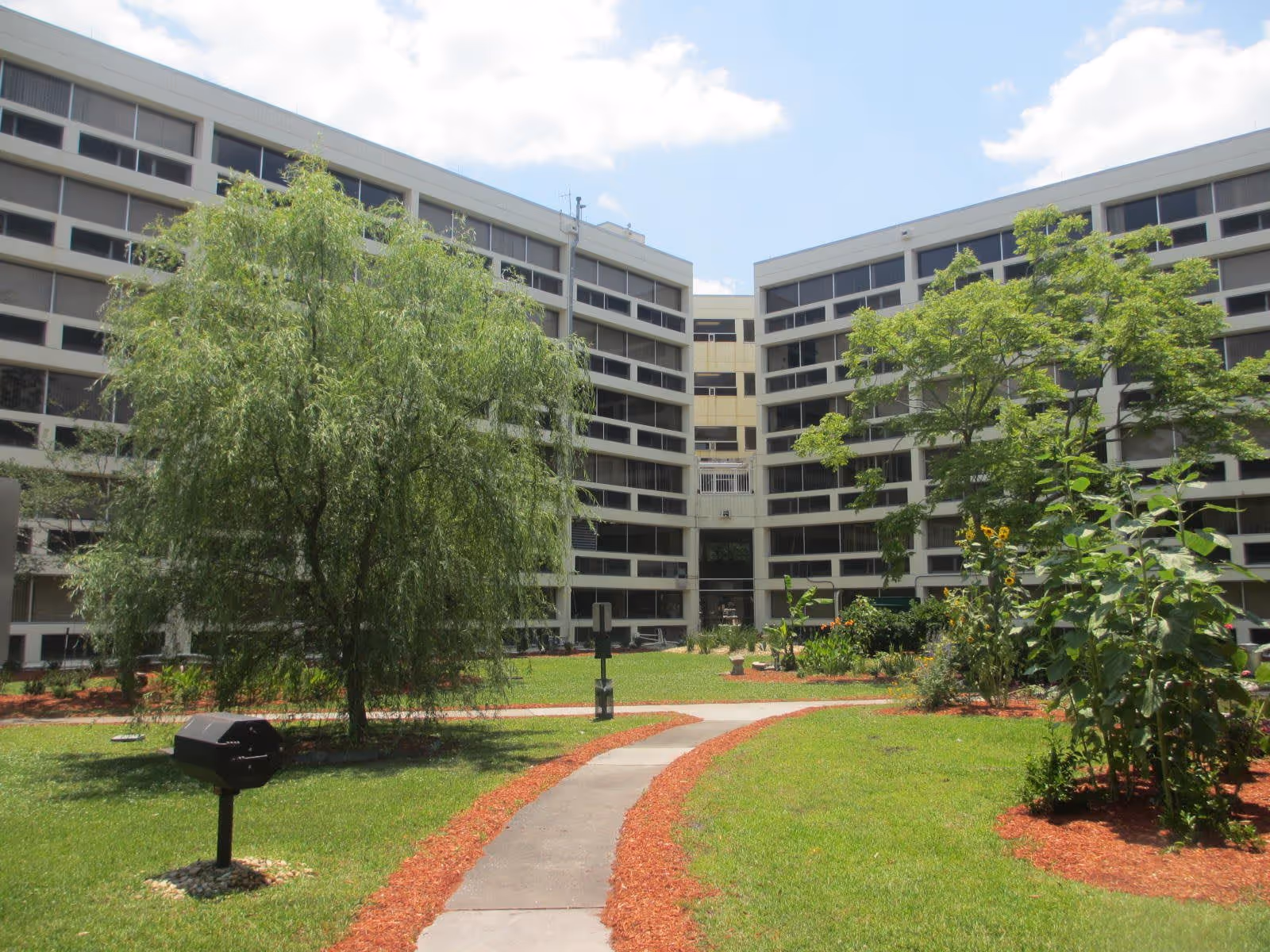 Outdoor courtyard area of Florida Christian Apartments featuring a paved walkway, green grass, various trees and plants, and a multi-story building with many windows in the background under a partly cloudy sky.