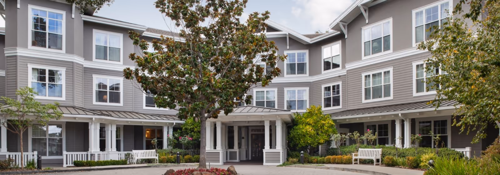 Exterior view of a three-story senior living facility building with gray siding and white trim. There is a covered entrance with white columns, benches, and landscaped greenery including trees and bushes in front of the building under a partly cloudy sky.
